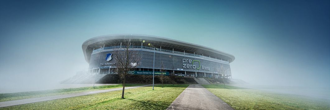 Die Außenansicht der PreZero Arena ist im Bildzentrum zu sehen. Teile des Geländes wie die Grünflächen und geteerten Wege zum Eingang des Stadions sind ebenfalls Teil des Bildes..