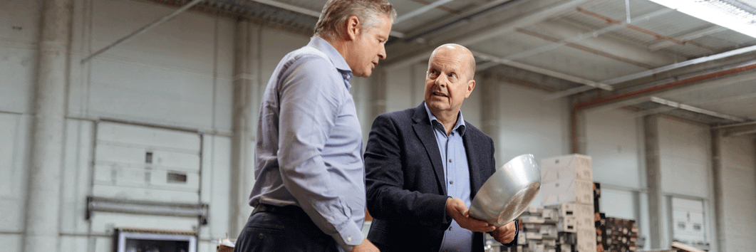 Zwei Männer in Business-Kleidung begutachten eine Metallschale in einer großen Industriehalle oder einem Lager