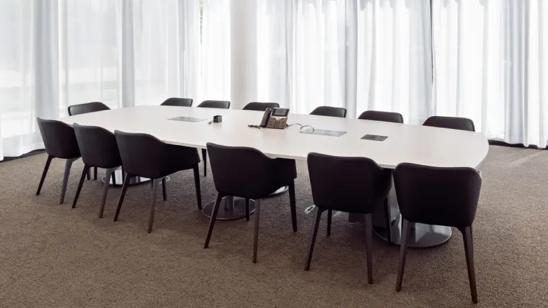Großer weißer Konferenzraumtisch mit schwarzen Polsterstühlen in einem hellen Raum mit Fensterfront