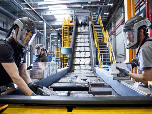 Arbeiter mit Gebläseatemschutz sortieren Batteriekomponenten an einem langen Förderband in einer Industriehalle.