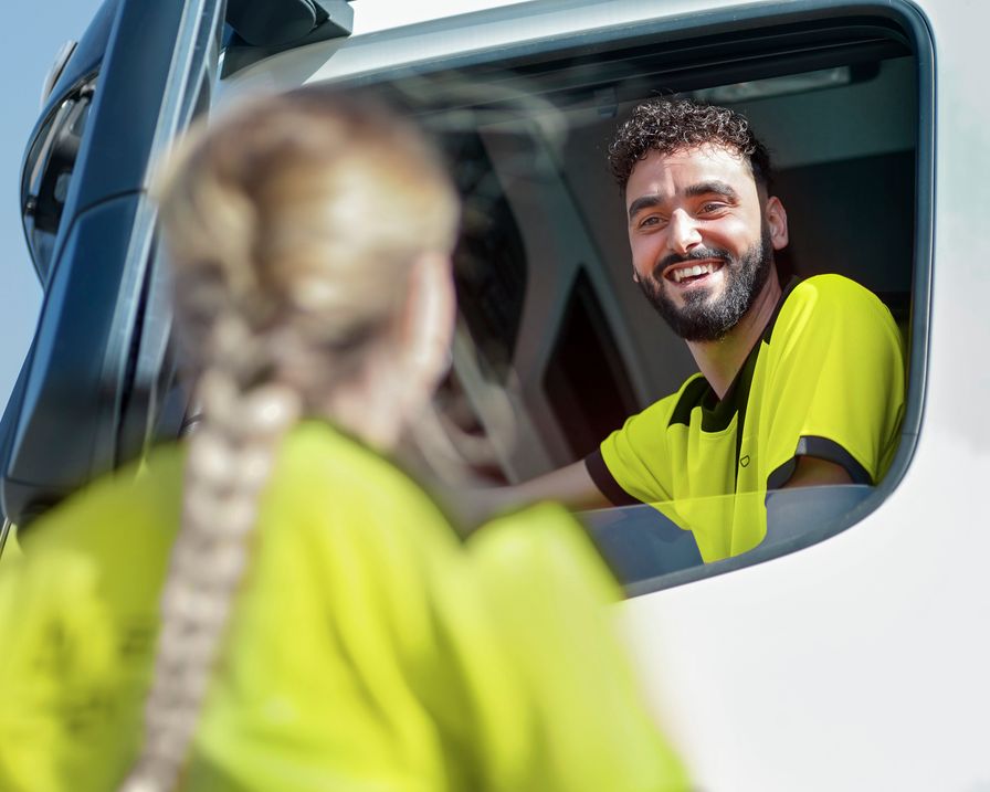 Ein lachender Lkw-Fahrer in gelber Warnkleidung blickt aus dem offenen Fenster seiner Fahrerkabine zu einer Kollegin, die unscharf im Vordergrund steht.
