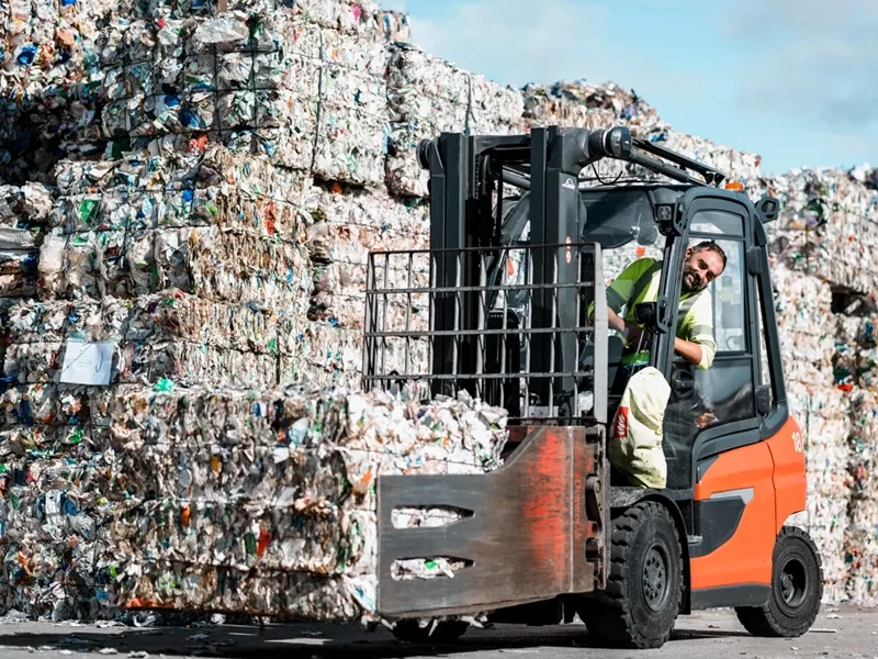 A worker drives a forklift carrying a pressed bale in front of huge stacks of waste at a plastic recycling facility