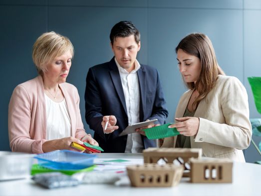 Drei Geschäftsleute begutachten bei einem Meeting am Tisch verschiedene Verpackungsmaterialien aus Plastik und Pappe.