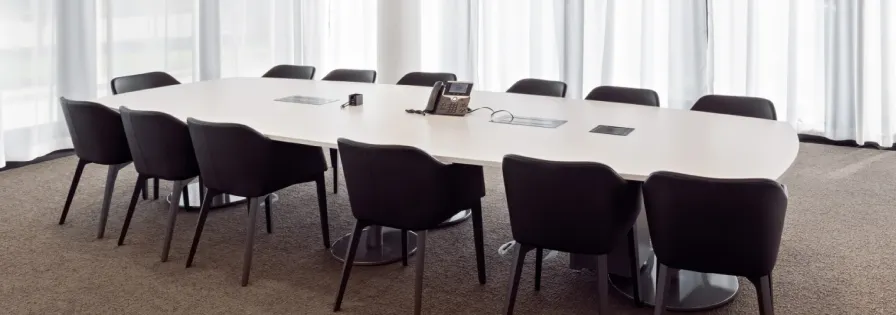 Großer weißer Konferenzraumtisch mit schwarzen Polsterstühlen in einem hellen Raum mit Fensterfront