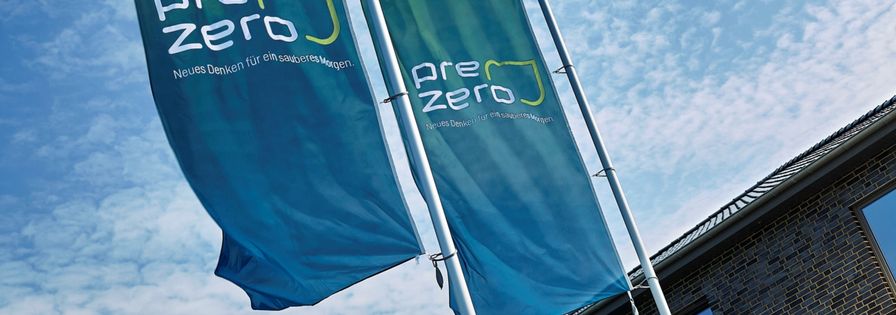 Two Bluegreen flags with the company name PreZero on it, a big brickhouse en a blue sky in the background