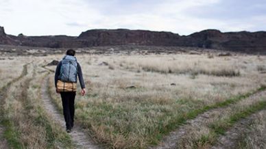 Man walking the left path after a fork in a plain, flat mountains in the background