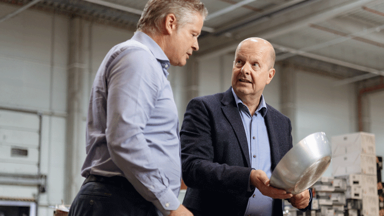 Zwei Männer in Business-Kleidung begutachten eine Metallschale in einer großen Industriehalle oder einem Lager