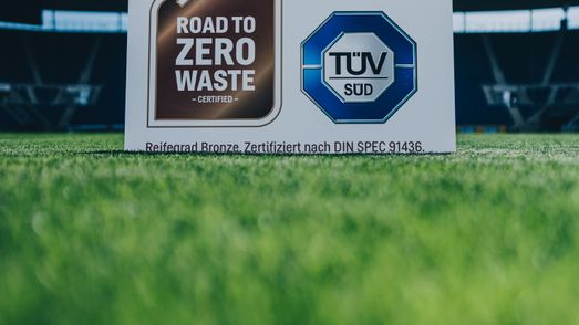 Zero-Waste-Zertifikat mit dem Reifegrad Bronze ist auf dem Rasen der Arena aufgestellt. Im Vordergrund sieht man den Rasen, im Hintergrund teile der Tribüne.
