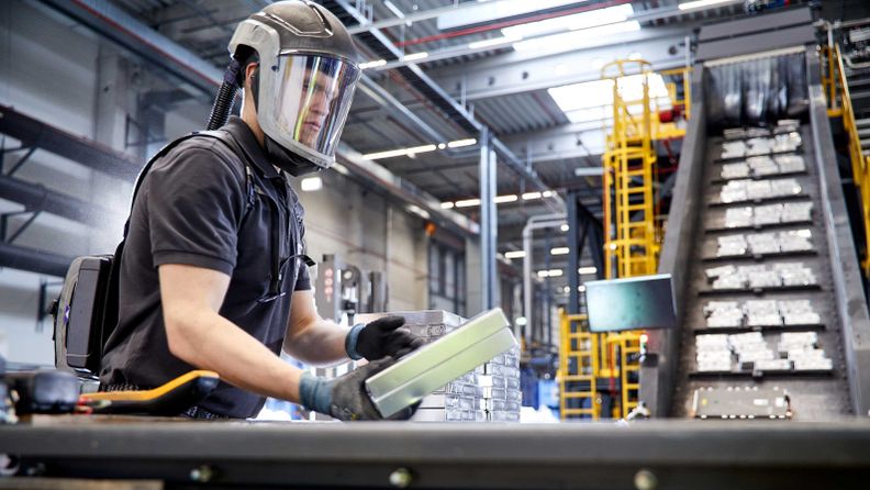 Arbeiter mit Gebläseatemschutz prüft eine silberne Batteriezelle an einer Sortieranlage in einer Industriehalle.