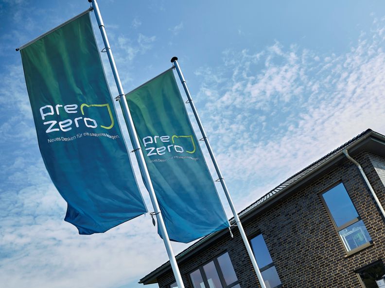 Two Bluegreen flags with the company name PreZero on it, a big brickhouse en a blue sky in the background