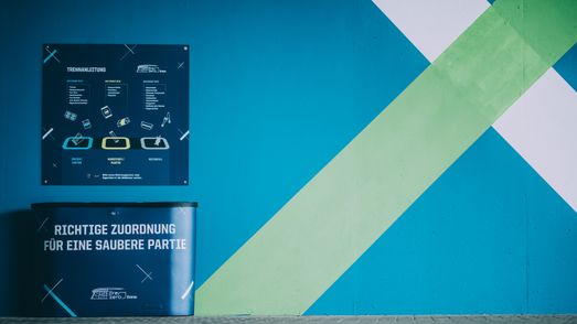 Ein Trennsystem vor einer Wand in der PreZero Arena ist links im Bild zu sehen. Über den dunkelblauen Mülleimern hängt eine Tafel, die Hinweise zur Abfalltrennung gibt.