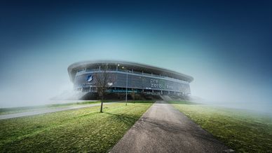 Die Außenansicht der PreZero Arena ist im Bildzentrum zu sehen. Teile des Geländes wie die Grünflächen und geteerten Wege zum Eingang des Stadions sind ebenfalls Teil des Bildes..