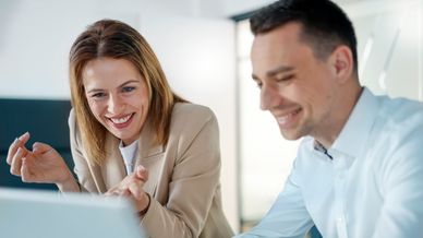Zwei lächelnde Mitarbeiter arbeiten gemeinsam am Laptop im Büro und unterhalten sich erfreut.