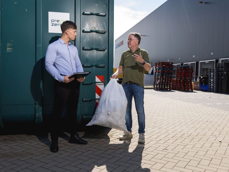 2 mannen staan voor groene containers te praten met een vuilniszak in de hand