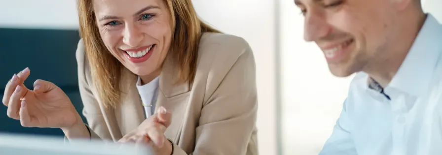 Eine Frau und ein Mann im Business-Outfit blicken lächelnd und gemeinsam auf einen Laptop-Bildschirm