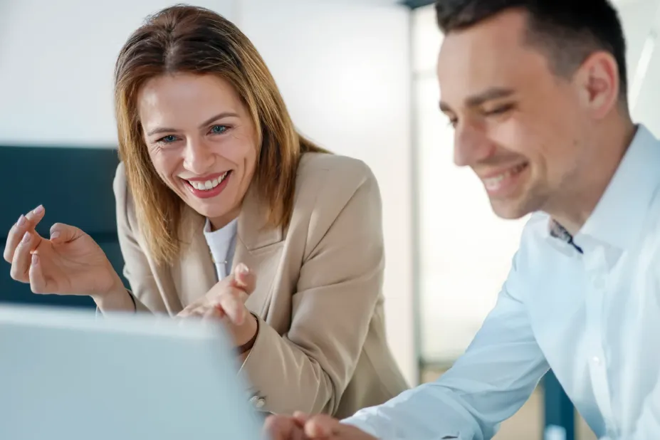 Eine Frau und ein Mann im Business-Outfit blicken lächelnd und gemeinsam auf einen Laptop-Bildschirm