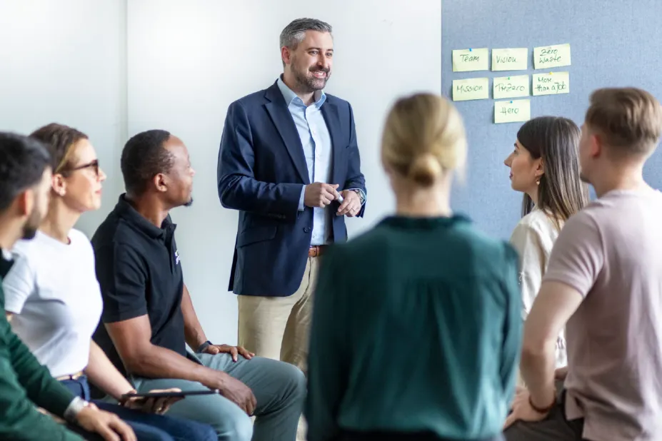 Ein Workshop-Leiter steht vor einer Gruppe von Kollegen, die im Halbkreis sitzen. Im Hintergrund hängen gelbe Notizzettel an einer grauen Pinnwand, auf denen Begriffe wie ‚Mission‘, ‚PreZero‘ und ‚Zero Waste‘ zu lesen sind