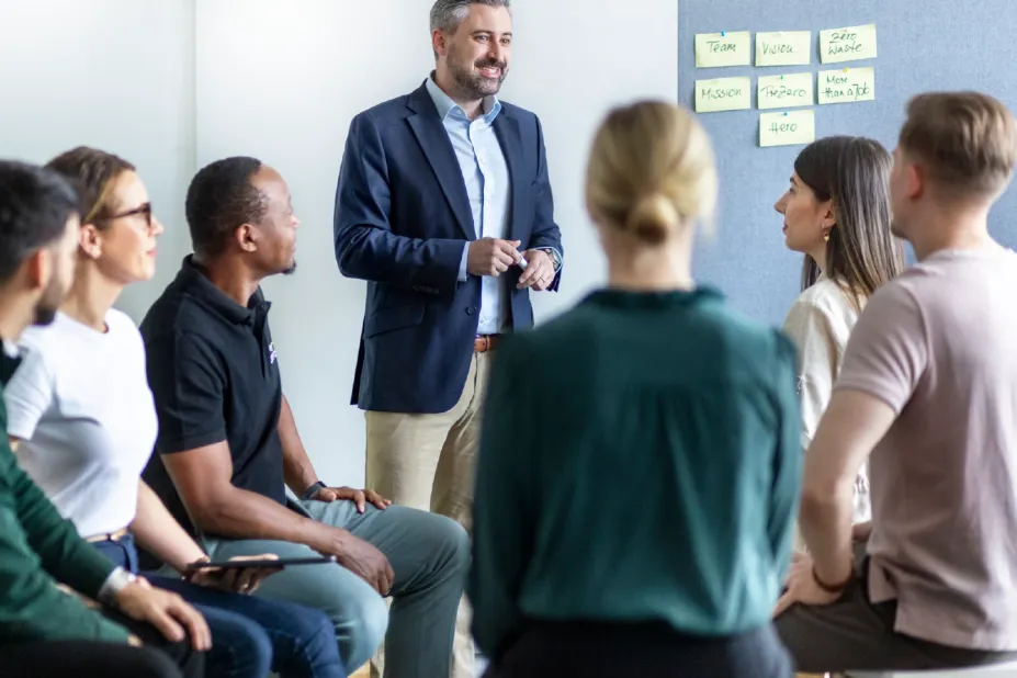 A male facilitator in a blue blazer and khakis stands before a seated semi-circle of five colleagues during a team-building event. Yellow sticky notes on a gray board list company values like 'Mission,' 'PreZero,' and 'Zero Waste