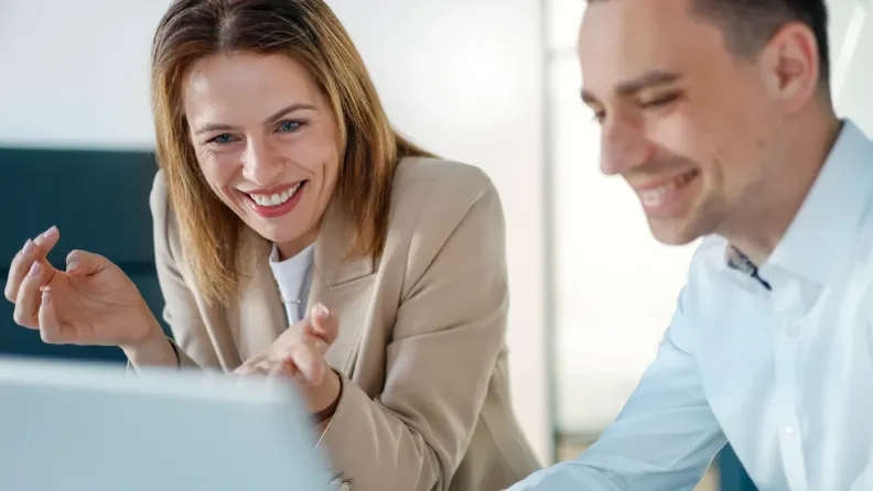 Eine Frau und ein Mann im Business-Outfit blicken lächelnd und gemeinsam auf einen Laptop-Bildschirm