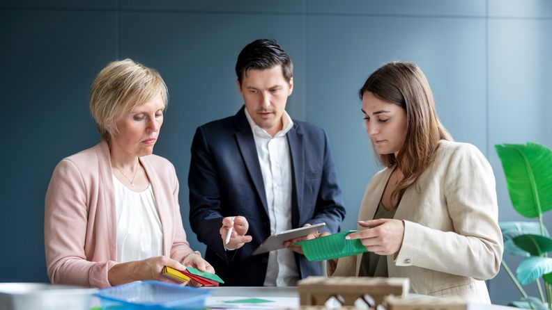 Drei Geschäftsleute begutachten bei einem Meeting am Tisch verschiedene Verpackungsmaterialien aus Plastik und Pappe.