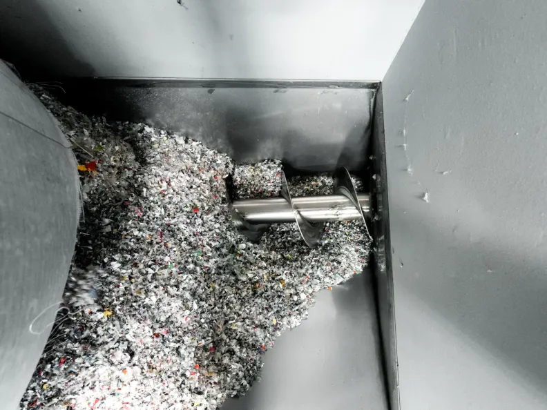 Analyse: Ein technischer Blick von oben in einen Trichter aus Edelstahl. Eine metallische Förderschnecke transportiert eine große Menge geschredderten Materials