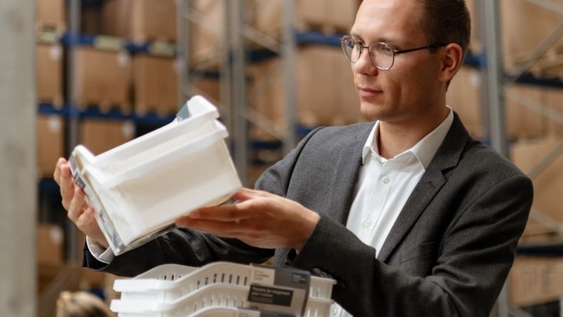 Ein Mann im Anzug begutachtet eine weiße Kunststoffbox in einem großen Hochregallager