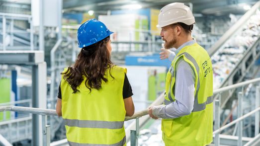 Zwei PreZero-Mitarbeiter in Warnwesten und Helmen besprechen Abläufe auf einem Steg in einer Recyclinganlage.