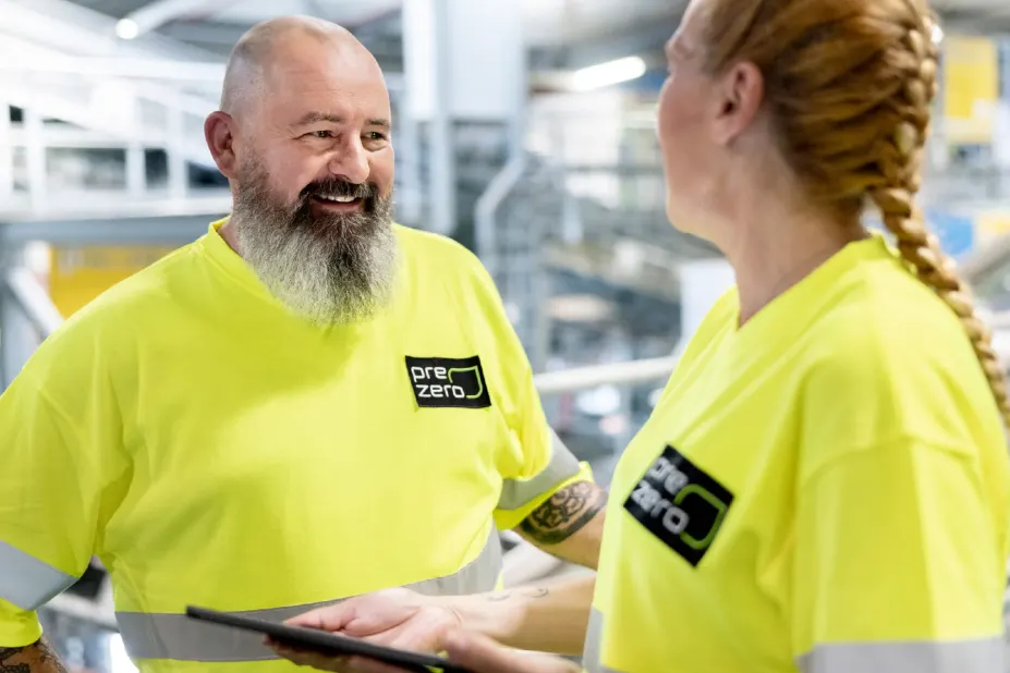 A man with a beard and a woman with braided hair wearing yellow PreZero safety shirts have a conversation in a warehouse or recycling plant while holding a tablet