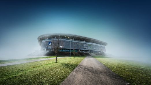Die Außenansicht der PreZero Arena ist im Bildzentrum zu sehen. Teile des Geländes wie die Grünflächen und geteerten Wege zum Eingang des Stadions sind ebenfalls Teil des Bildes..