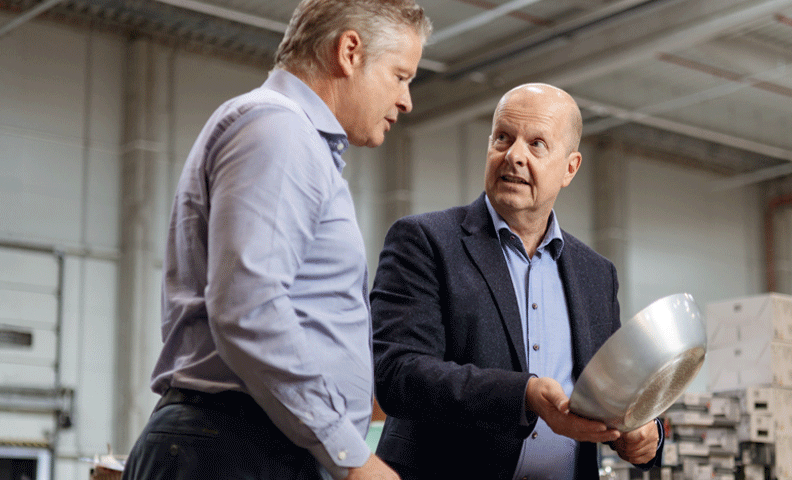 Zwei Männer in Business-Kleidung begutachten eine Metallschale in einer großen Industriehalle oder einem Lager