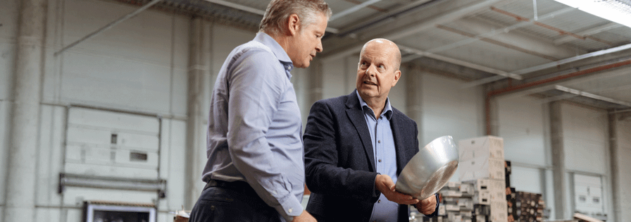 Zwei Männer in Business-Kleidung begutachten eine Metallschale in einer großen Industriehalle oder einem Lager