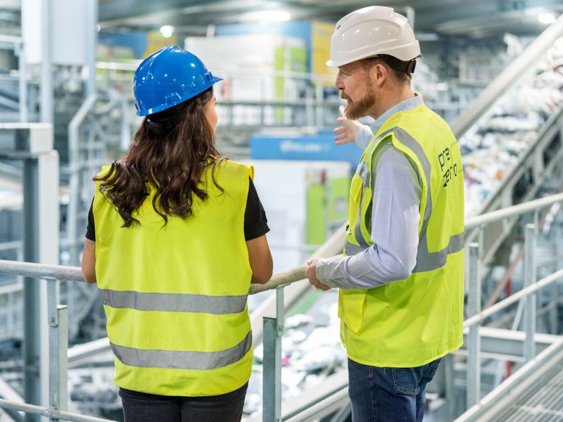 Zwei PreZero-Mitarbeiter in Warnwesten und Helmen besprechen Abläufe auf einem Steg in einer Recyclinganlage.
