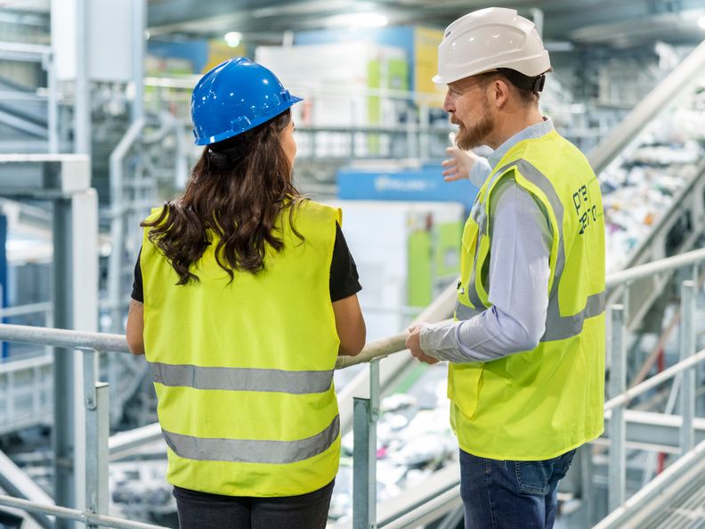 Zwei PreZero-Mitarbeiter in Warnwesten und Helmen besprechen Abläufe auf einem Steg in einer Recyclinganlage.